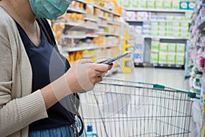 Young woman shopping in supermarket with trolley. Female hands using smart phone in store. Woman wearing medical mask ptotection