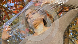 Young woman with short blonde hair is looking through a plastic bag, polyethylene wrap standing outdoors durind sunny
