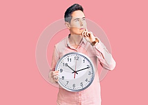 Young woman with short hair holding big clock serious face thinking about question with hand on chin, thoughtful about confusing