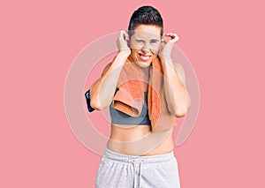 Young woman with short hair wearing sportswear and towel using smartphone covering ears with fingers with annoyed expression for
