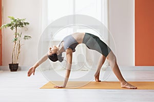A young woman in short leggings and a T-shirt, does yoga, performs in the room, standing on the mat, kamatkarasana exercise
