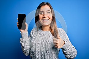 Young woman showing blank smartphone screen over blue isolated background happy with big smile doing ok sign, thumb up with