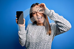 Young woman showing blank smartphone screen over blue isolated background with happy face smiling doing ok sign with hand on eye