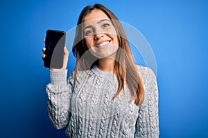 Young woman showing blank smartphone screen over blue isolated background with a happy face standing and smiling with a confident