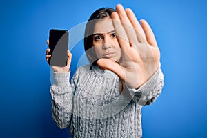 Young woman showing blank smartphone screen over blue isolated background with open hand doing stop sign with serious and