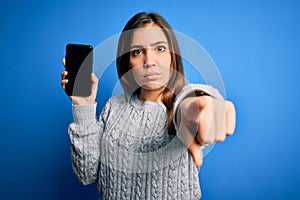 Young woman showing blank smartphone screen over blue isolated background pointing with finger to the camera and to you, hand