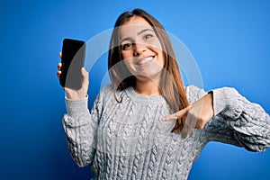 Young woman showing blank smartphone screen over blue isolated background with surprise face pointing finger to himself