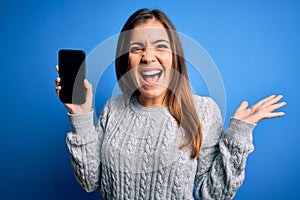 Young woman showing blank smartphone screen over blue isolated background very happy and excited, winner expression celebrating