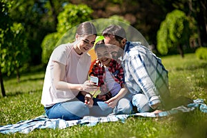 Young Woman Is Showing Photos On Phone To Her Husband And Daughter While Sitting On The Blanket In The Park.