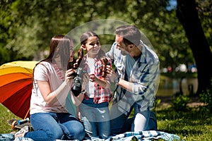 Young Woman Showing Photos To Her Husband And Her Daughter While Sitting On Blanket In The Park.