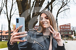 Young woman showing victory sign during video call on smart phone at city park.woman holding smartphone and showing V victory sign