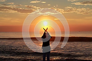 A young woman silhouette raising her hands towards the sky and standing on the sea shore at sunrise in the morning, welcoming and