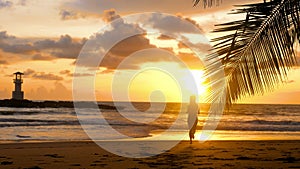 Young woman silhouette running barefoot on sandy beach to ocean with sunset sun, orange sky and lighthouse on background