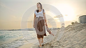 Young woman with a silk scarf walking at sunset on a sandy sea beach. Happiness, travel, and holiday concepts