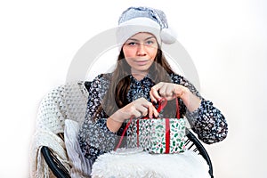 Young woman in a silver santa hat sitting on a chair and holding a gift box in her hands, isolated on a white background