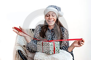 Young woman in a silver santa hat sitting on a chair and holding a gift box in her hands, isolated on a white background