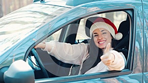 Young woman sit in car during travelling break. Happy positive girl wear red Santa hat and smile. Hold big thumbs up