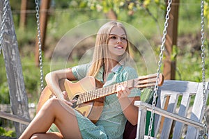 Young Woman sit on swing and playing guitar in summer park. Female model with guitar. Girl playing guitar outdoor