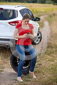 Young woman siting next to broken car and searching for auto service number in mobile phone