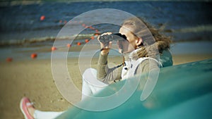 Young woman sits on the beach outside of the boat and looks through binoculars