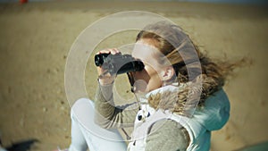 Young woman sits on the beach outside of the boat and looks through binoculars