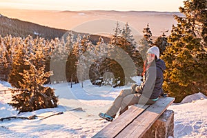 A young woman sits on a bench in the mountains and rests after hiking on a winter mountain trail