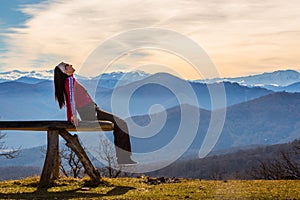 Young woman sits on bench outside and look at pictorial landscape with mountains