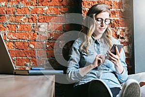 Young woman sits in cafe on windowsill and uses smartphone. Hipster girl checking e-mail, chatting, blogging, working.