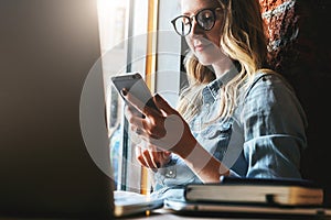 Young woman sits in cafe on windowsill and uses smartphone. Hipster girl checking e-mail, chatting, blogging, working.