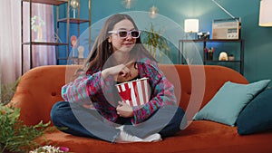 Young woman sits on couch eating popcorn, watching interesting TV serial sport game online at home