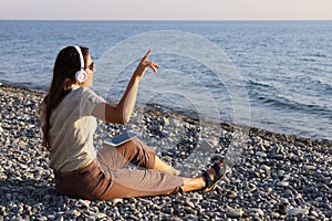 Young woman sits on deserted seashore listening to music and singing along to the rhythm