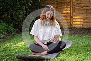 A young woman sits on a fitness mat and looks at the smartphone screen.