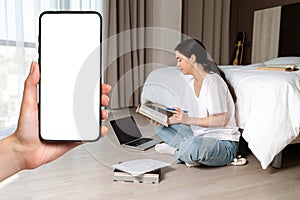 A young woman sits on the floor and reading a book. There are papers and a laptop on the floor. A close-up hand holding