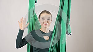 Young woman sits on a hammock smiling and waving her hand, a greeting gesture, hello, hi. Aerial aero fly fitness
