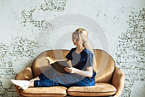 A young woman sits at home on a beige sofa on a background of a white brick wall and reads a book. Self-development and home