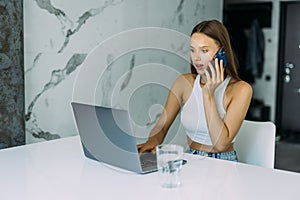 Young woman sits at the kitchen table using a laptop and talking on a ce phone