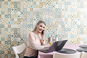 Young woman sits at the kitchen table using a laptop and talking on a cell phone