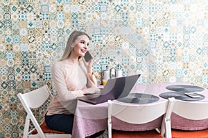 Young woman sits at the kitchen table using a laptop and talking on a cell phone