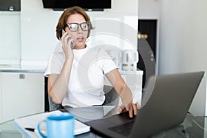 Young woman sits at the kitchen table using a laptop and talking on a cell phone