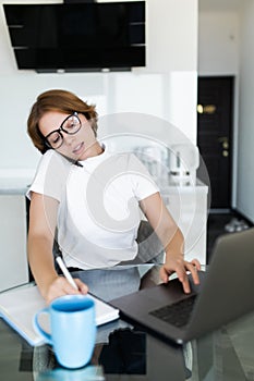 Young woman sits at the kitchen table using a laptop and talking on a cell phone