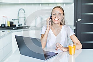 Young woman sits at the kitchen table using a laptop and talking on a cell phone