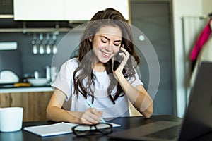 Young woman sits at the kitchen table using a laptop and talking on a cell phone