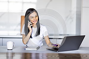 Young woman sits at the kitchen table using a laptop and talking on a phone