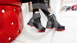 Young woman sits in a mall on a soft chair in stylish black jeans in fashionable black leather seasonal red boots. Autumn-spring