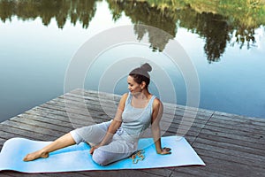 A young woman sits on a mat in a free pose by the lake. Yoga class in nature