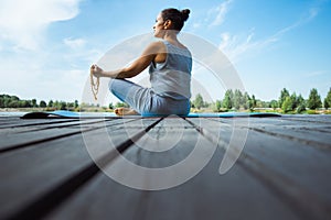 A young woman sits on a mat on a wooden pier in a lotus position with a rosary in her hands. Back view. Against the background of
