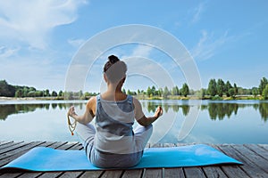 A young woman sits on a mat on a wooden pier in a lotus position with a rosary in her hands. Back view. Meditation, yoga in nature