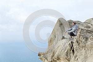 A young woman sits on a mountain top high above the sea on a sunny day. Cute blonde girl in a sports jacket and jeans. Active