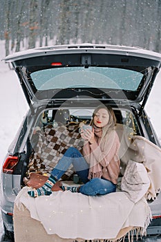 Young woman sits with a mug of coffee squinting in pleasure in the trunk of a car in a winter forest