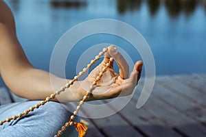 A young woman sits on the pier in a lotus position holding a rosary in her hand. Legs view. Meditation, yoga in nature. Close-up